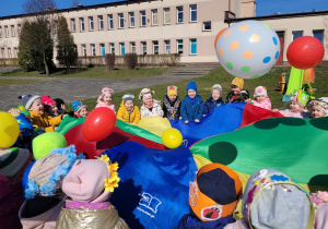 Dzieci uczestniczą w zabawie z chustą animacyjną i podrzucają kolorowe balony.