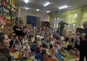 Dzieci ze wszystkich grup siedzą na dywanie słuchając Pani nauczycielki