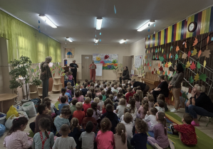 Dzieci ze wszystkich grup siedzą na dywanie słuchając Pani nauczycielki