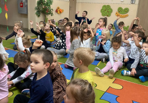 Dzieci siedzą na dywanie i poruszają rękoma w rytm granego utworu.