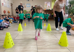Dzieci w zielonych koszulkach przenoszą kasztany na łyżeczkach.