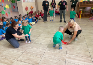 Przedszkolaki w zielonych koszulkach z pomocą swoich pań przechodzą przez szarfy.