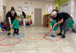 Chłopcy w zielonych koszulkach z pomocą pań przesuwają za pomocą kijka obręcze.
