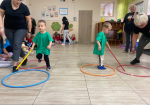 Chłopcy w zielonych koszulkach z pomocą pań przesuwają za pomocą kijka obręcze.