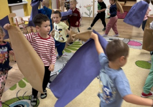 Dzieci naśladują odgłosy wiatru za pomocą dużych arkuszy papieru.