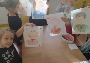Dzieci przy stoliku pokazują narysowane portrety babć.
