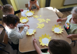 Dzieci pracują przy stolikach: przyklejają płatki do papierowego talerzyka