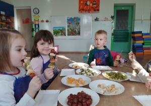 Dzieci siedzą przy stoliku i pokazują wykonane szaszłyki owocowe