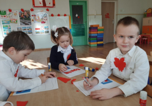 Dzieci siedzą przy stoliku i wyklejają wydzieranym papierem flagi