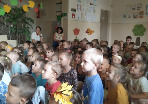 W holu przedszkolnym na kolorowym dywanie siedzi grupa dzieci oraz dwie nauczycielki. Oglądają przedstawienie. Atmosfera jest radosna.