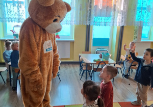W sali przedszkolnej na kolorowym dywanie stoi osoba przebrana za Misia Teddiego Eddiego. Przed nim stoi chłopczyk i dziewczynka. Wyglądają jakby do niego mówili. Atmosfera jest radosna.