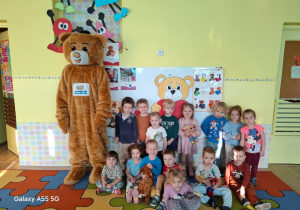 W sali przedszkolnej na kolorowym dywanie osoba przebrana za misia oraz grupa dzieci ustawiły się do zdjęcia. Atmosfera jest radosna.